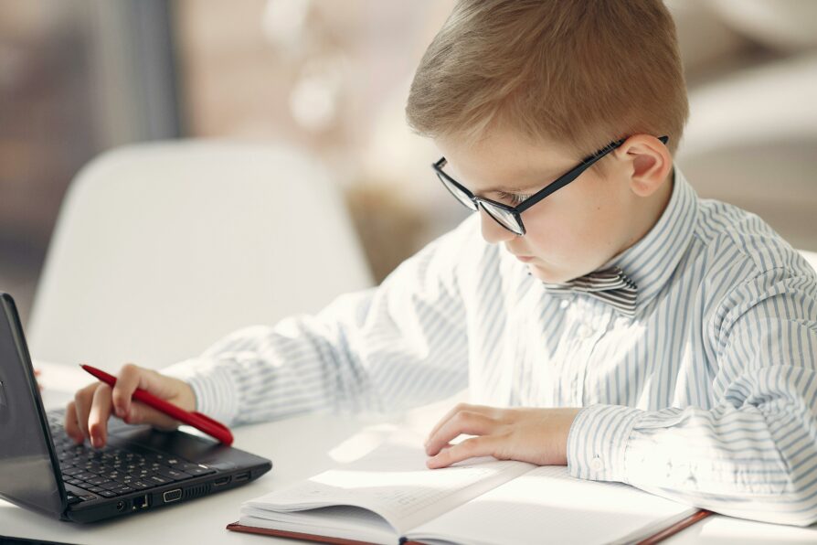 young kid wearing a button up shirt and tie doing homework and using his laptop